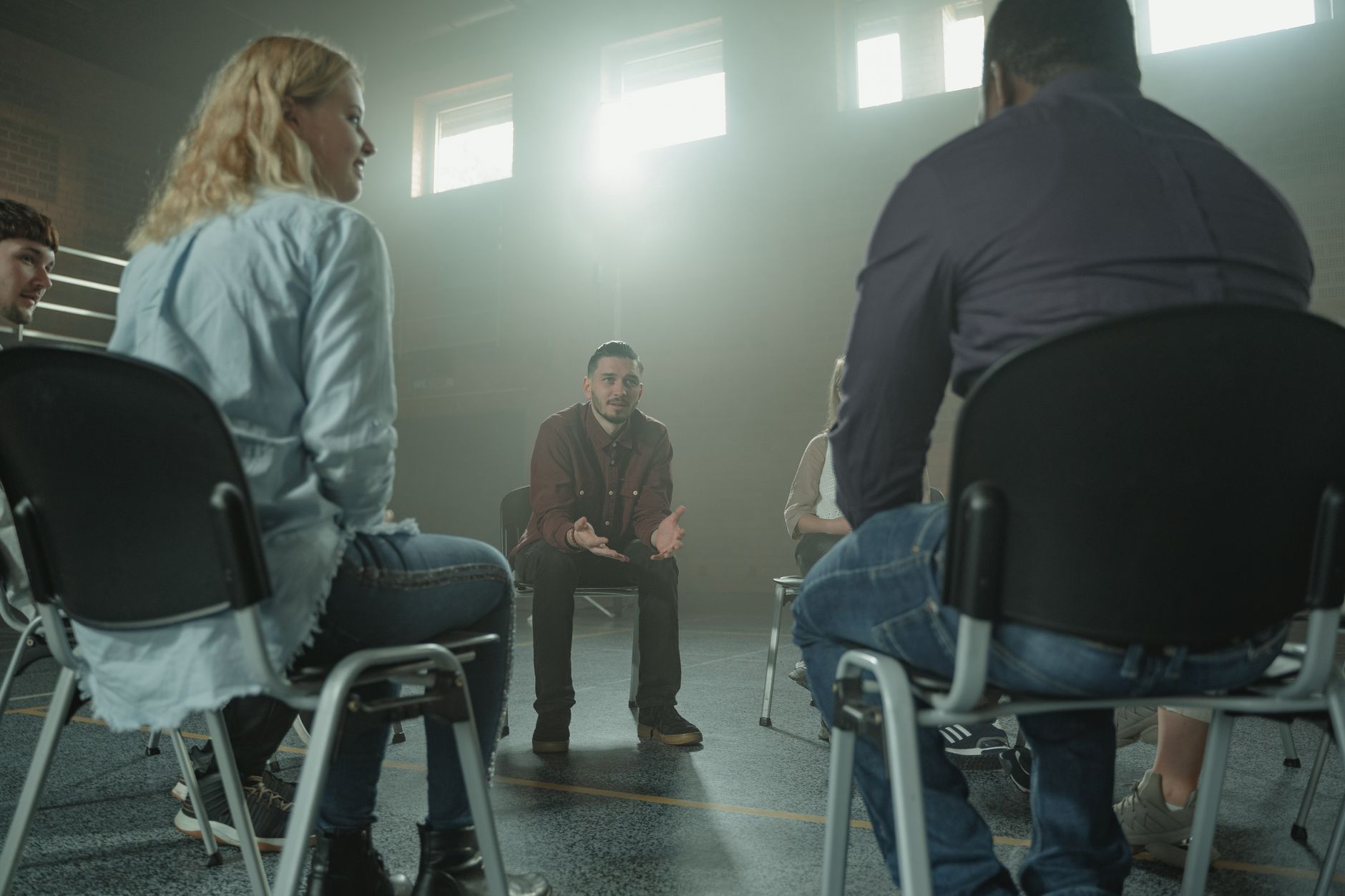 Adults sit in a circle of chairs in a sunlit hall, listening as one man speaks