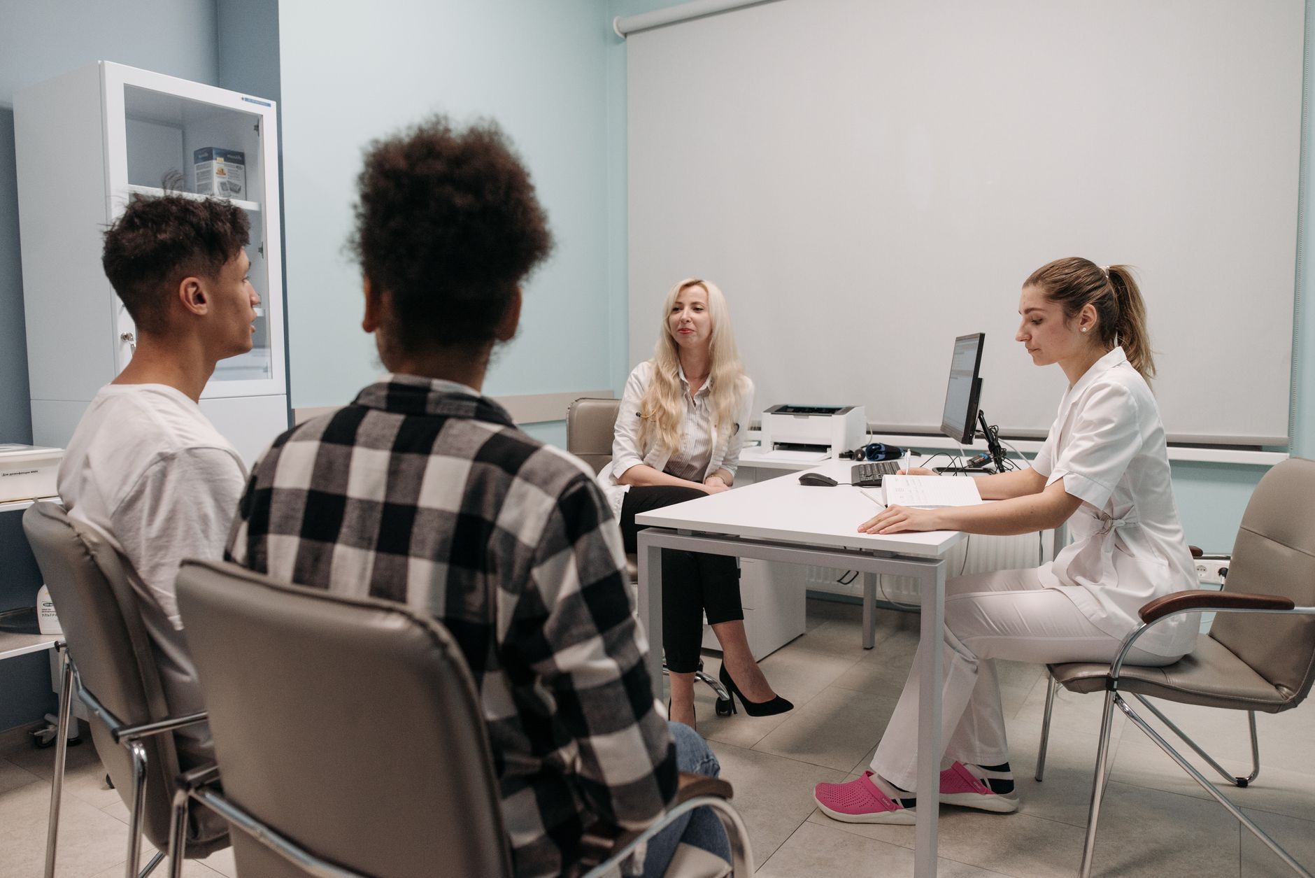 Two young adults sit across a desk from two clinicians in a bright clinic office, talking through a first appointment