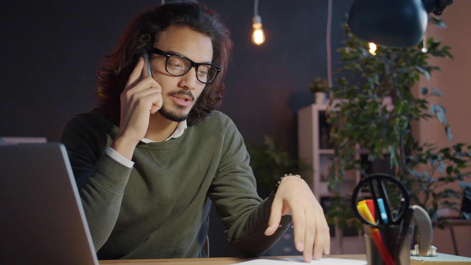 A man in glasses talks on his phone at a desk with a laptop and notes, as if calling about his benefits