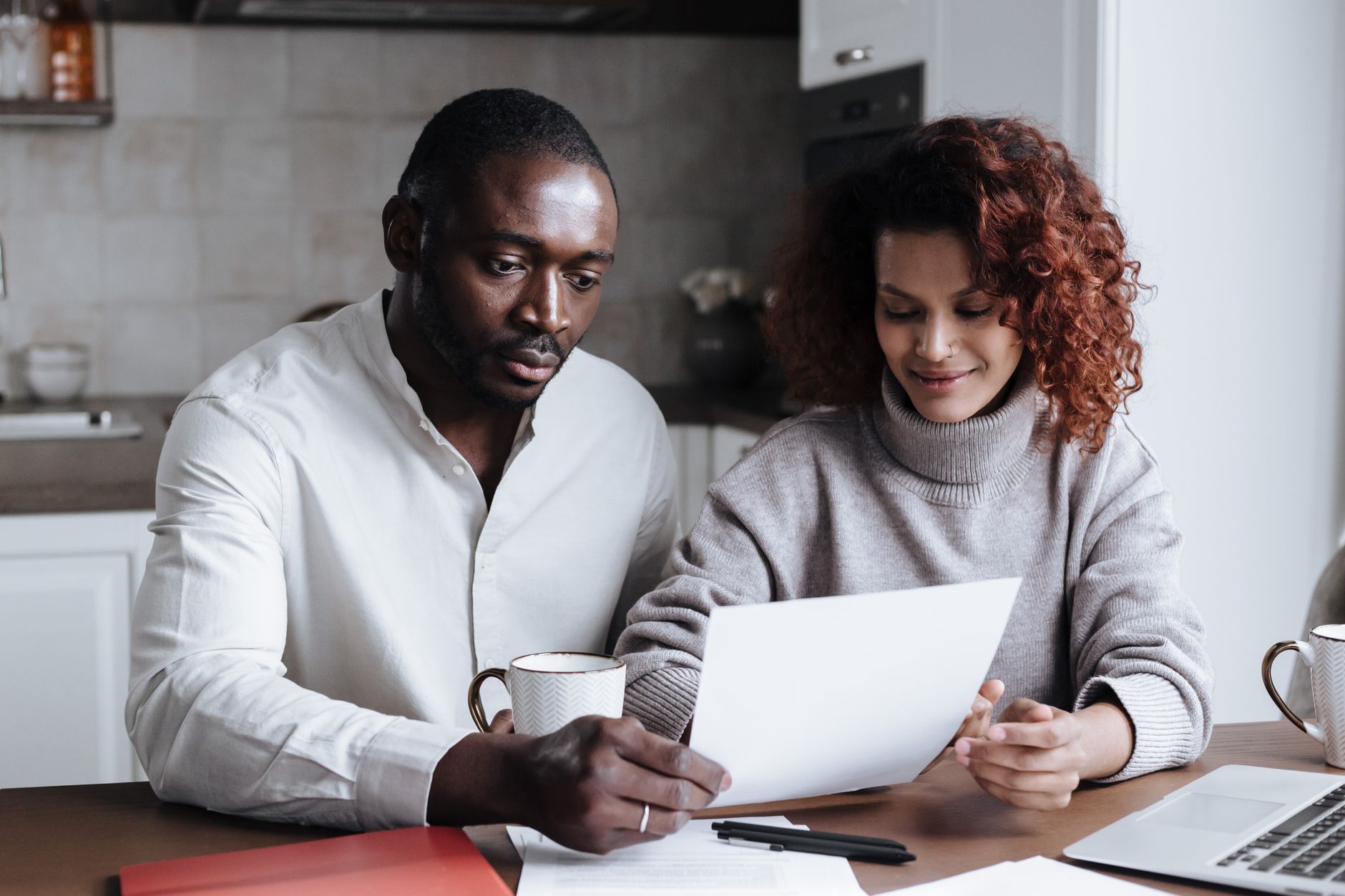 A couple sits at a kitchen table with coffee mugs and a laptop, reading a sheet of paper together