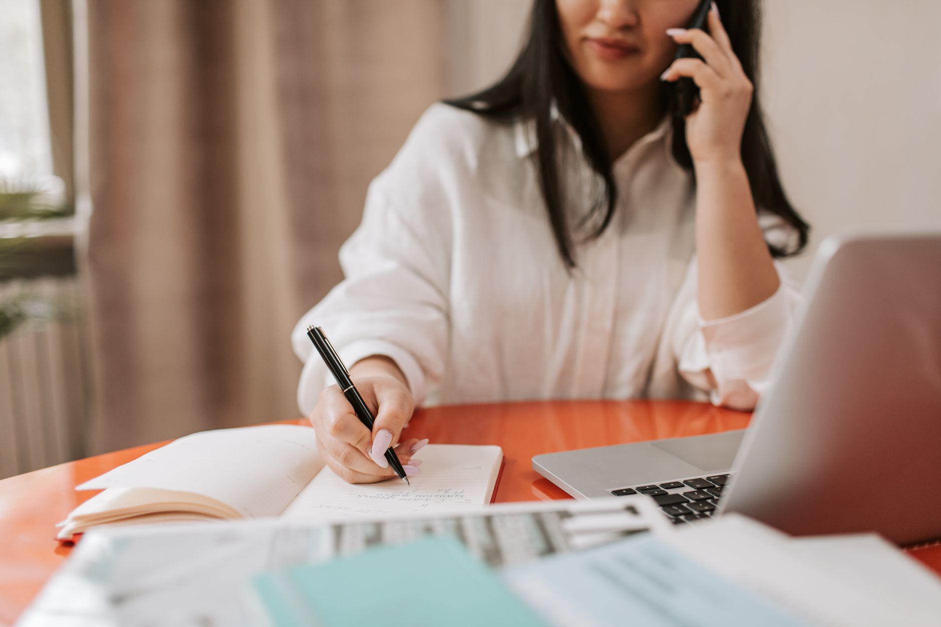 A woman on a phone call writes notes in a notebook at a table beside a laptop