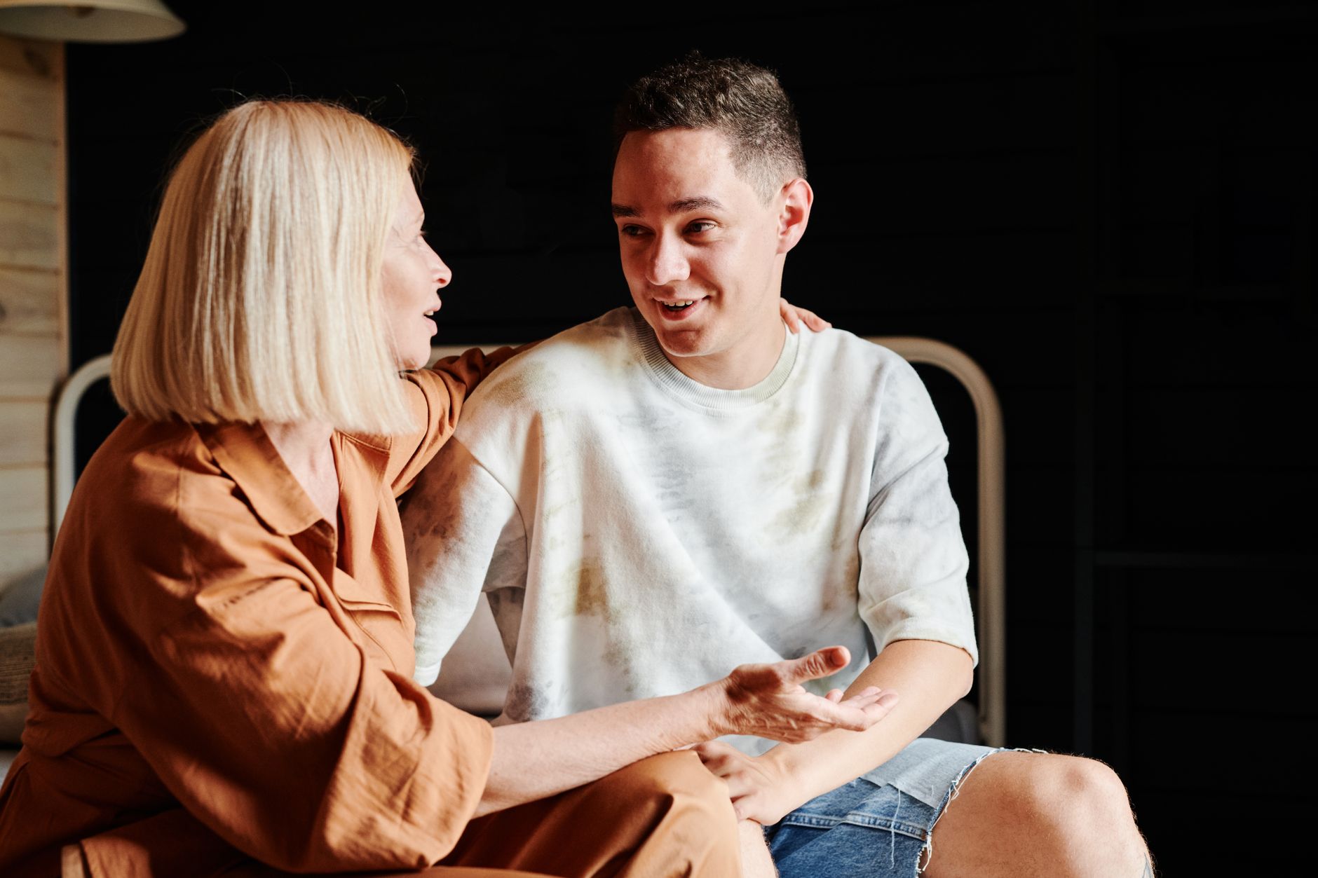 An older woman rests her hand on a young man's shoulder as they sit side by side and talk