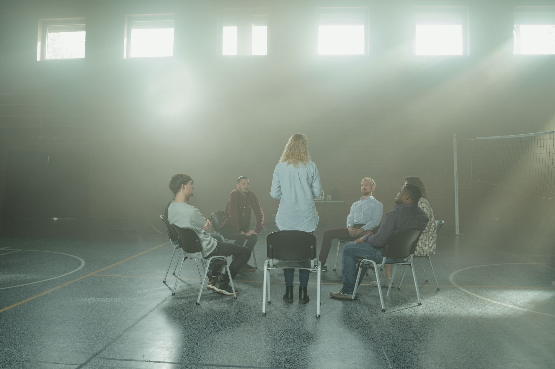 A small group of adults sit on chairs in a circle in a sunlit hall while a facilitator stands and talks with them