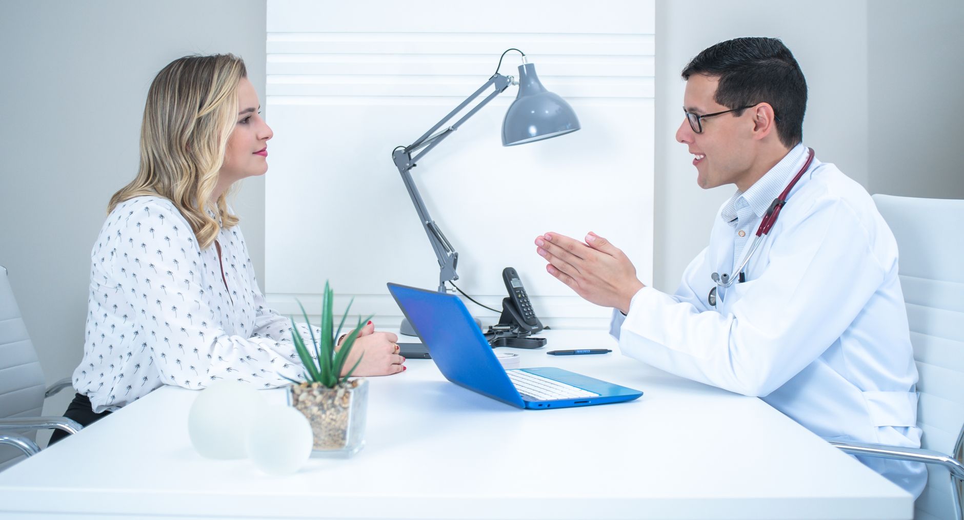 A doctor in a white coat talks with a woman across a bright office desk