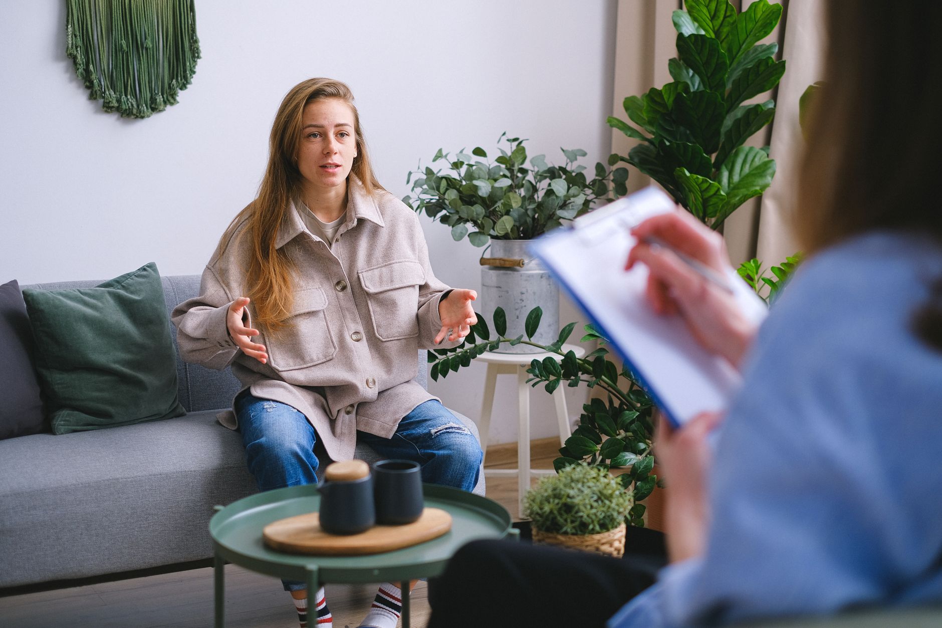 A young woman sits on a sofa and talks while a counselor across from her takes notes in a calm, plant-filled room