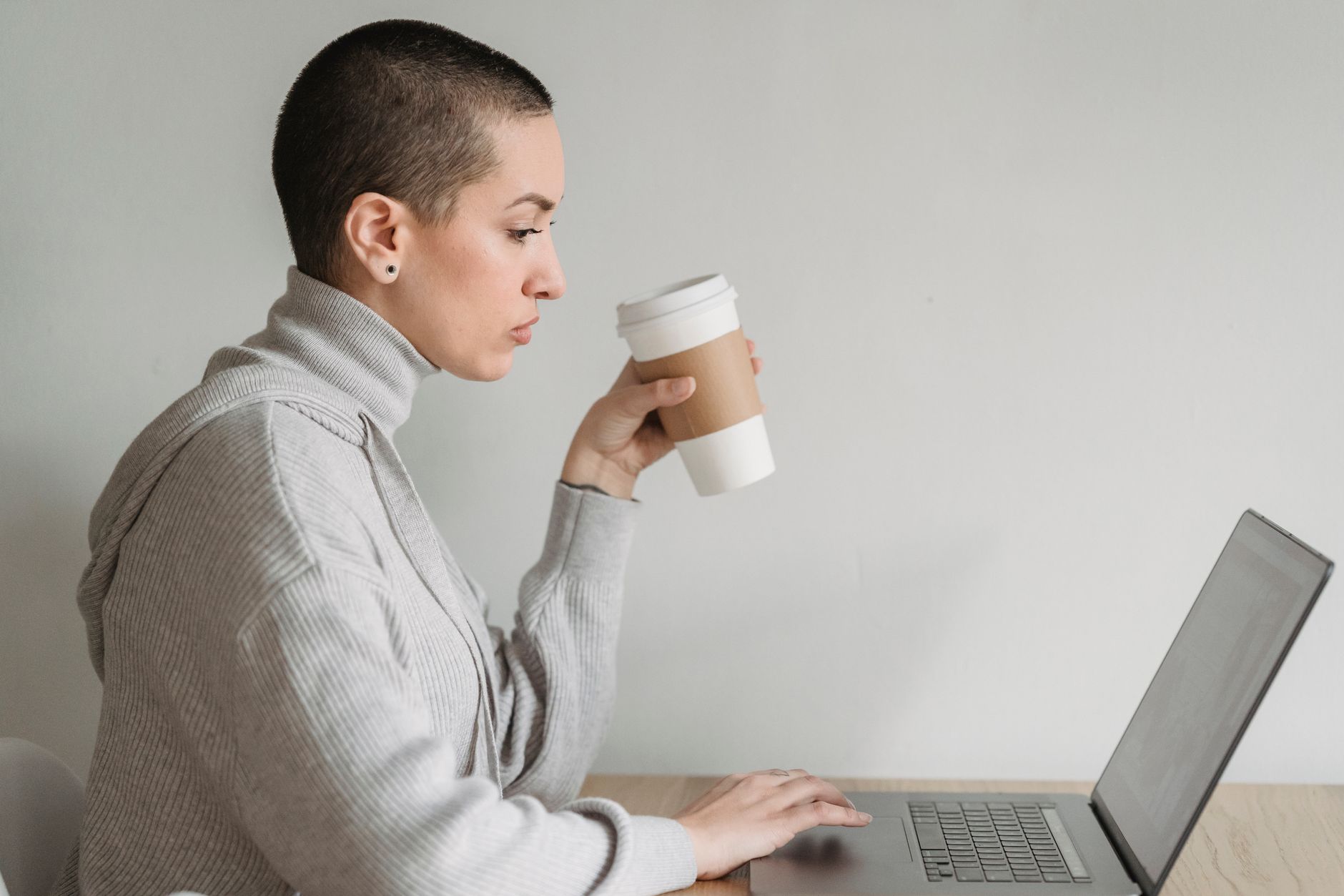A woman with short hair sips from a paper cup while working on a laptop at a table