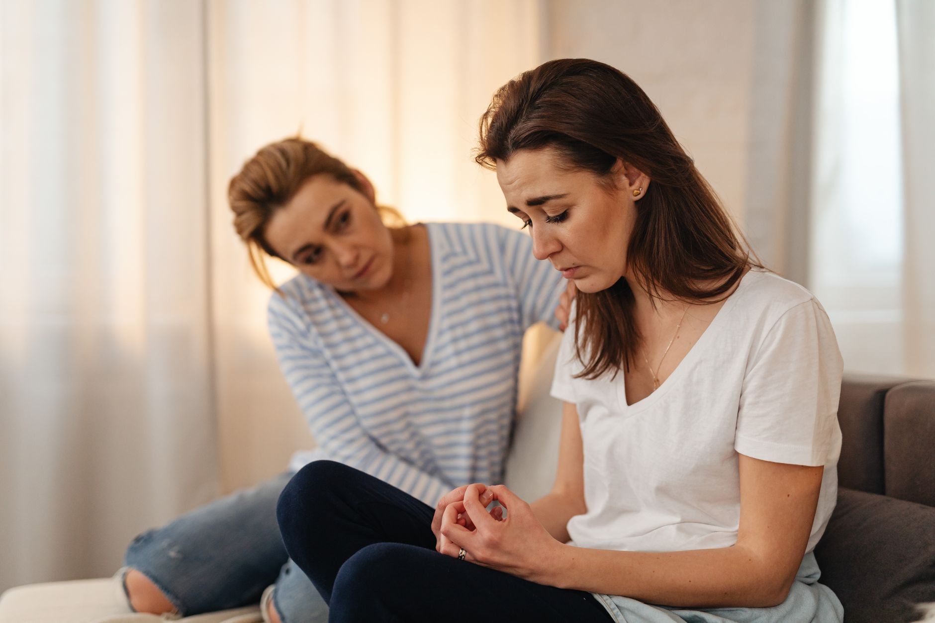 A woman sits on a sofa with her hand on the shoulder of a friend who looks down, worried