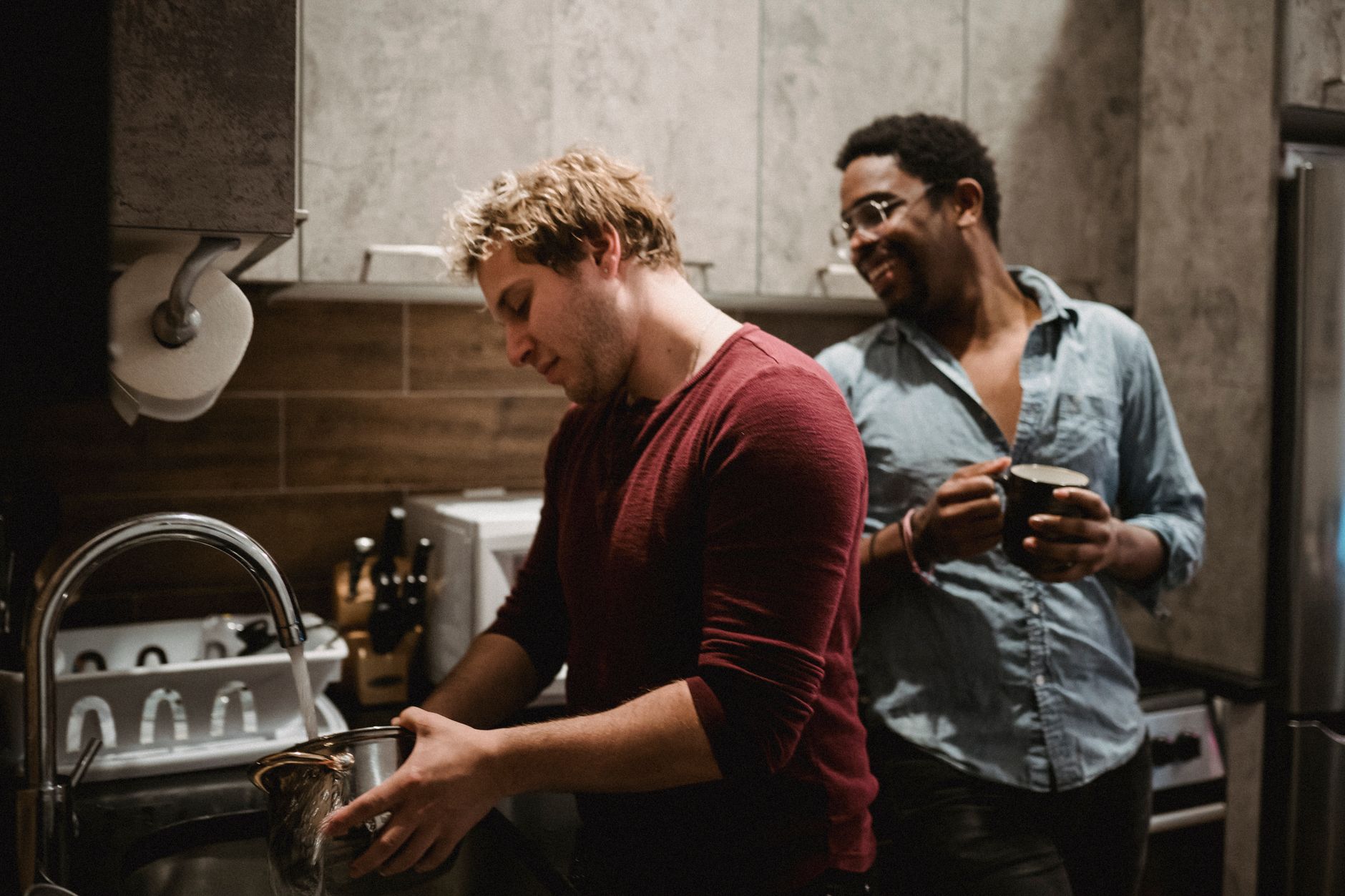 Two men share a kitchen, one rinsing a pot at the sink while the other holds a mug and smiles