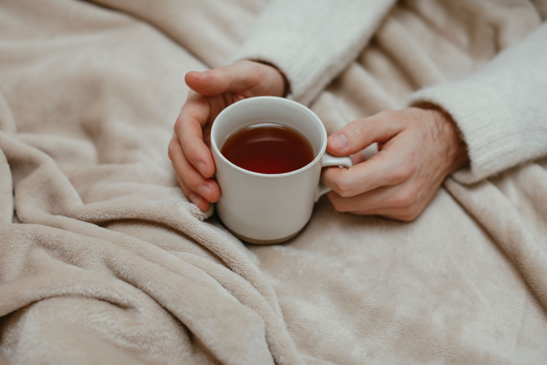 Two hands wrapped around a white mug of tea on a soft beige blanket